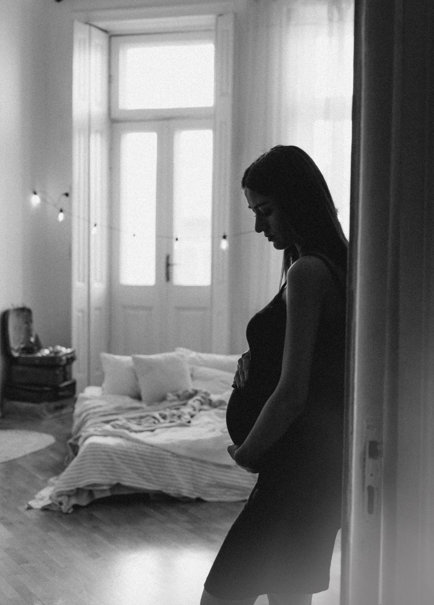 A pregnant woman in a bedroom doorway at dusk, both hands resting on her belly, looking down