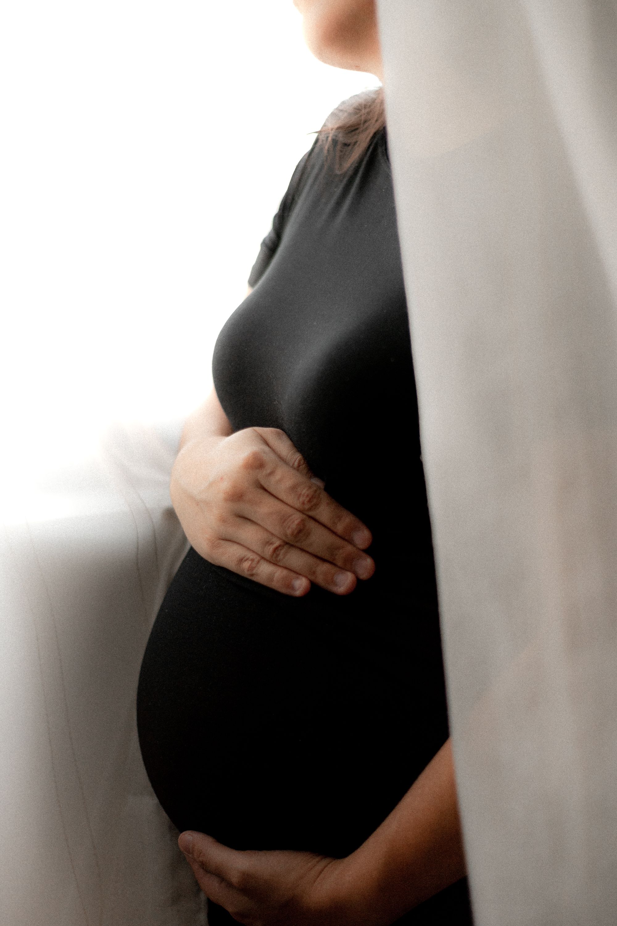 A pregnant woman in a simple dress by a soft-lit window, both hands cradling her belly